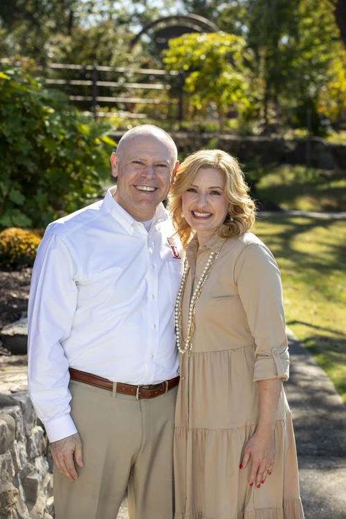 President Brad Johnson is standing beside his wife in a warm, elegant portrait. He is wearing a crisp white button-down shirt, while his wife is dressed in a soft beige sweater accented by a beautiful gold-yellow necklace. Their coordinated, neutral-colored clothing creates a polished and timeless appearance, with the necklace adding a subtle touch of warmth and sophistication.